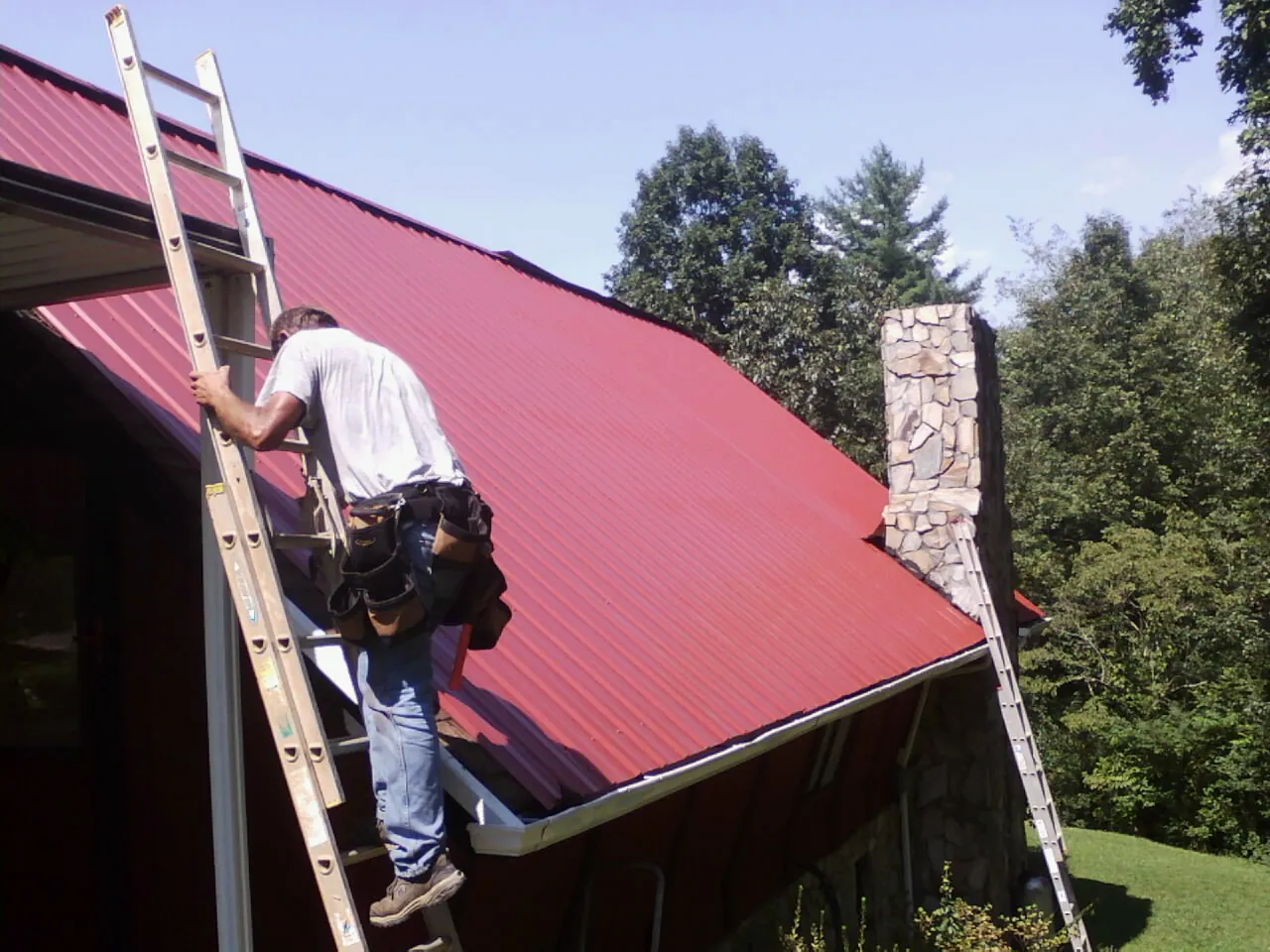 Roofer on a ladder beside a red metal roof and stone chimney — Soffit & Fascia Repair by Roofing Brothers in Paragon